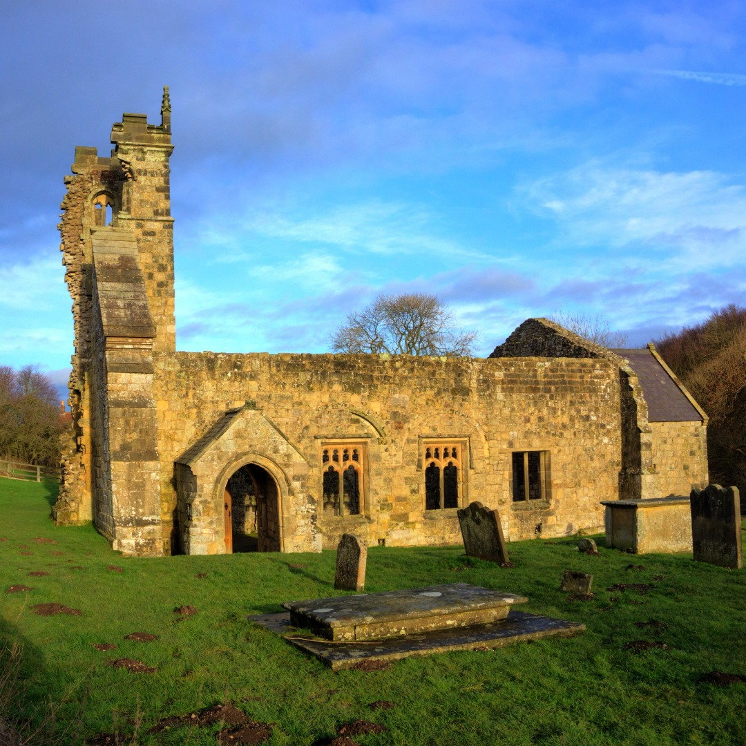 A photo of the church ruins of Wharram Percy, surrounded by green grass, autumn trees, and a blue sky in the early evening.